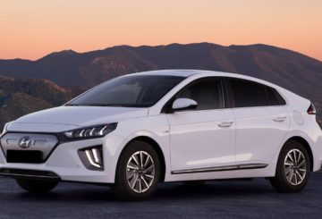 Foto de um Hyundai Ioniq 2022. O carro é um sedã branco com rodas estilizadas e detalhes em prata e preto. Ele está estacionado em uma rua de asfalto e atrás dele é possível ver montanhas e o pôr-do-sol