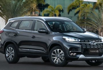 Foto de um Chery Tiggo 8, ano modelos 2021, na cor cinza. Na foto, o carro está estacionado em frente a um prédio que tem palmeiras na frente.