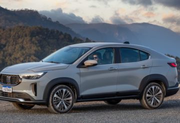 Foto de um Fiat Fastback 2023 na cor cinza. Na foto é possível ver uma parte da dianteira e a lateral esquerda do carro, que está estacionado em uma estrada de asfalto. Atrás é possível ver uma montanha coberta por nuvens.