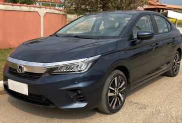 Foto de um Honda City 2022, na versão Touring, cinza escuro. O Carro é sedan e tem um detalhe cromado que atravessa quase toda a tampa do capô e ele está estacionado em uma rua de asfalto, em frente a um muro cor de salmão com detalhes em branco.
