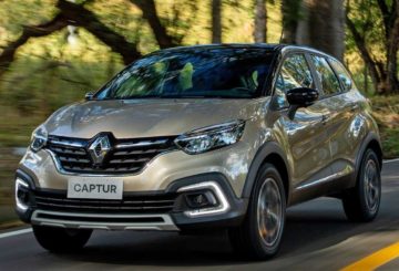 Foto de um Renault Captur 2022 na cor cinza amarelado. Na foto, o carro está sendo dirigido em uma estrada e, ao fundo, é possível ver árvores.