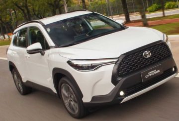 Foto de um Toyota Corolla Cross branco, sendo dirigido em uma rua de asfalto. Ao fundo é possível ver a calçada e um canteiro com árvores.