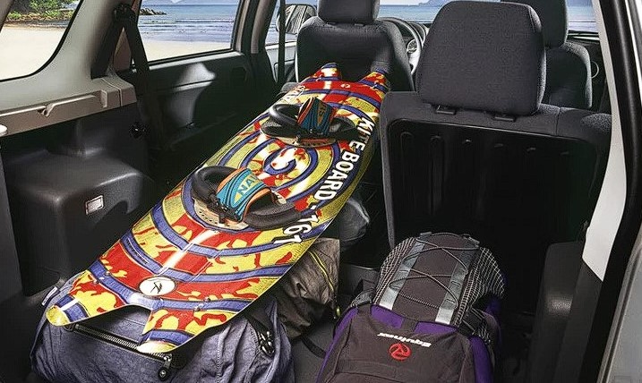 Foto mostrando o espaço do porta-malas do Ecosport G1. Na foto é possível ver as costas dos bancos traseiros, sendo que um deles está abaixado, para acomodar uma prancha e 3 mochilas.