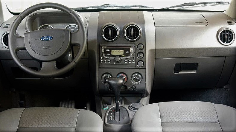 Foto do interior do carro. Nela, é possível ver o volante, o painel, os controles e as saídas de ar, o porta-luvas e os assentos dos bancos dianteiros. O interior é preto com detalhes em cinza.