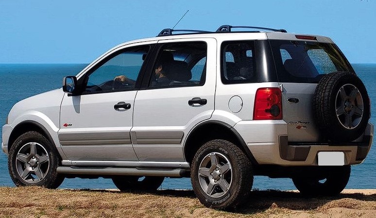 Foto da traseira de um Ford Ecosport de primeira geração. Na foto, o carro está estacionado na grama e ao fundo é possível ver o mar e o céu azul.