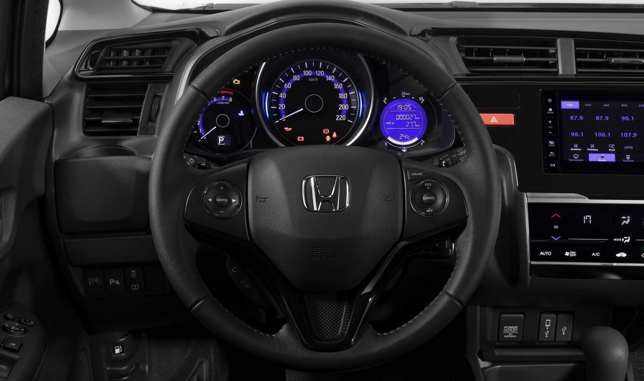 Foto do interior do carro mostrando o volante, o velocímetro, as saídas de ar do lado do motorista e uma parte da central multimídia. O ambiente é predominantemente preto, com luzes de LED que variam entre vermelho, azul e branco. Os detalhes dos botões também são brancos.