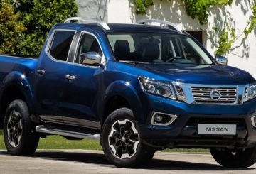 Foto da dianteira de um Nissan Frontier G3 azul. Na foto, o carro está estacionado em uma rua de asfalto e, ao fundo, é possível ver as paredes brancas de uma casa e alguns arbustos verdes.