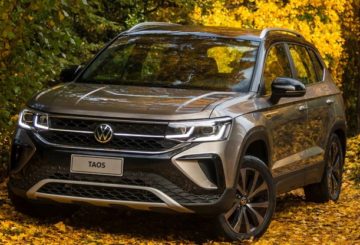 Foto de um Volkswagen Taos prateado, estacionado de frente em um lugar cheio de árvores com folhas verdes e amarelas espalhadas pelo chão.