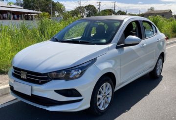 Foto de um Fiat Cronos branco estacionado de frente em uma rua de asfalto. Ao fundo, é possível ver a calçada, um canteiro com grama verde alta e o céu azul claro.