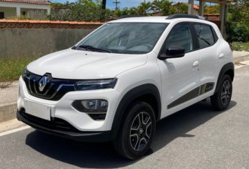 Foto de um Renault Kwid E-Tech branco estacionado de frente em uma rua de asfalto. Ao fundo é possível ver a calçada, um canteiro de grama e o muro de concreto de uma casa.