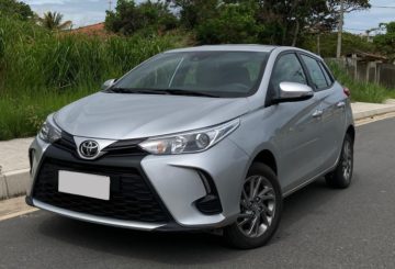 Foto de um Toyota Yaris 2023 prateado estacionado de frente em uma rua de asfalto. Ao fundo, é possível ver a calçada e um canteiro com algumas árvores verdes.