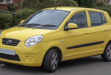 Foto mostrando a dianteira de um Kia Picanto G1 amarelo que está estacionado em uma rua de asfalto.
