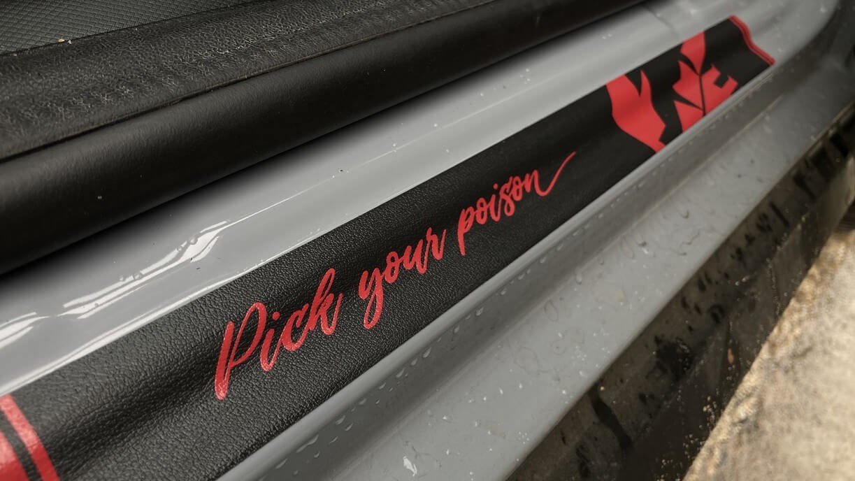 Foto de detalhe mostrando uma etiqueta em couro preto com detalhes em vermelho que fica na parte de baixo da abertura da porta do motorista. Nela está escrito a frase "Pick your poison" em letra cursiva vermelha.