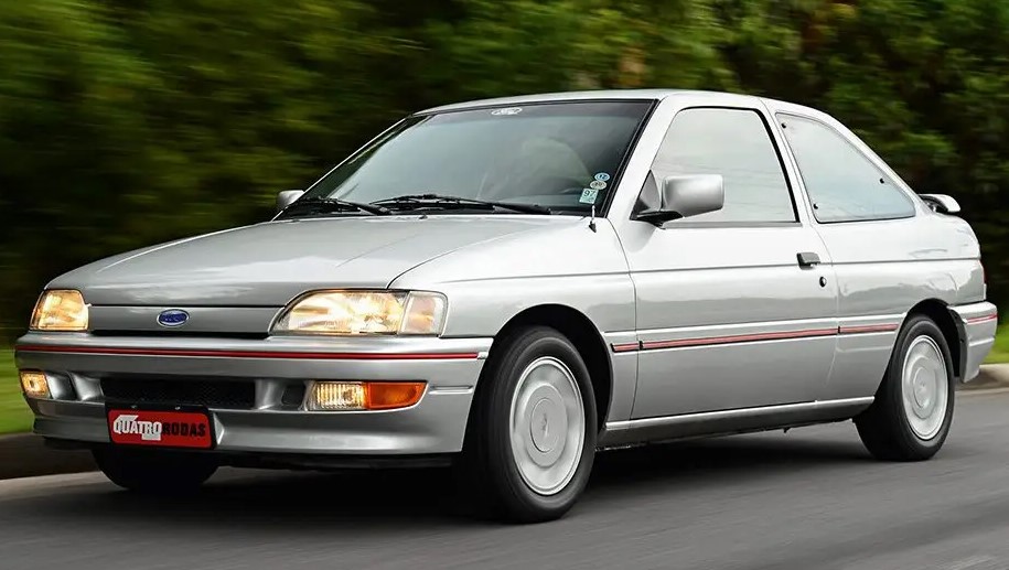Foto mostrando um Ford Escort 1993, versão XR3, prateado sendo dirigido em uma estrada de asfalto.