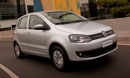 Foto mostrando a diagonal direita de um VW Fox 2013 prateado que está sendo dirigido em uma rua de asfalto.