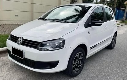 Foto mostrando a diagonal esquerda de um Volkswagen Fox 2014 branco que está parado em frente ao muro de uma casa.