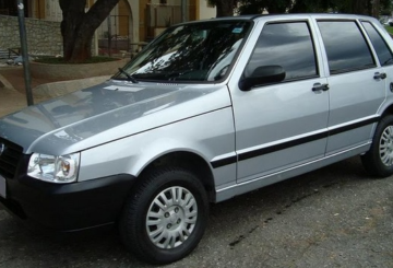 Foto mostrando a diagonal esquerda de um Fiat Uno Mille 2006 prateado estacionado em uma rua de asfalto.