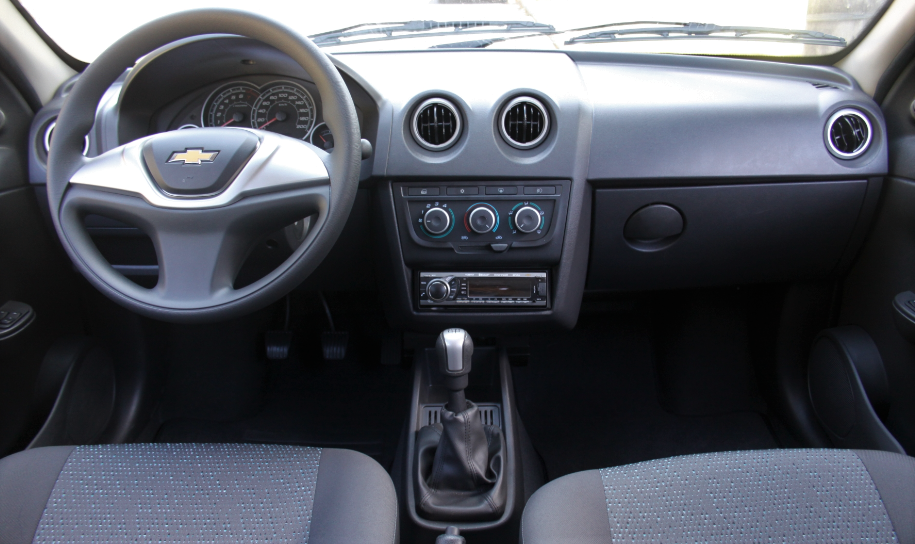Foto da cabine do carro. É possível ver o volante, o painel, as saídas e os comandos de ar e do rádio, o câmbio e os assentos dos bancos dianteiros.