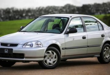 Foto mostrando a diagonal esquerda de um Honda Civic G6 prateado estacionado em uma rua de asfalto.