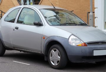Foto mostrando a diagonal esquerda de um Ford Ka G1 prateado estacionado em uma rua de asfalto.