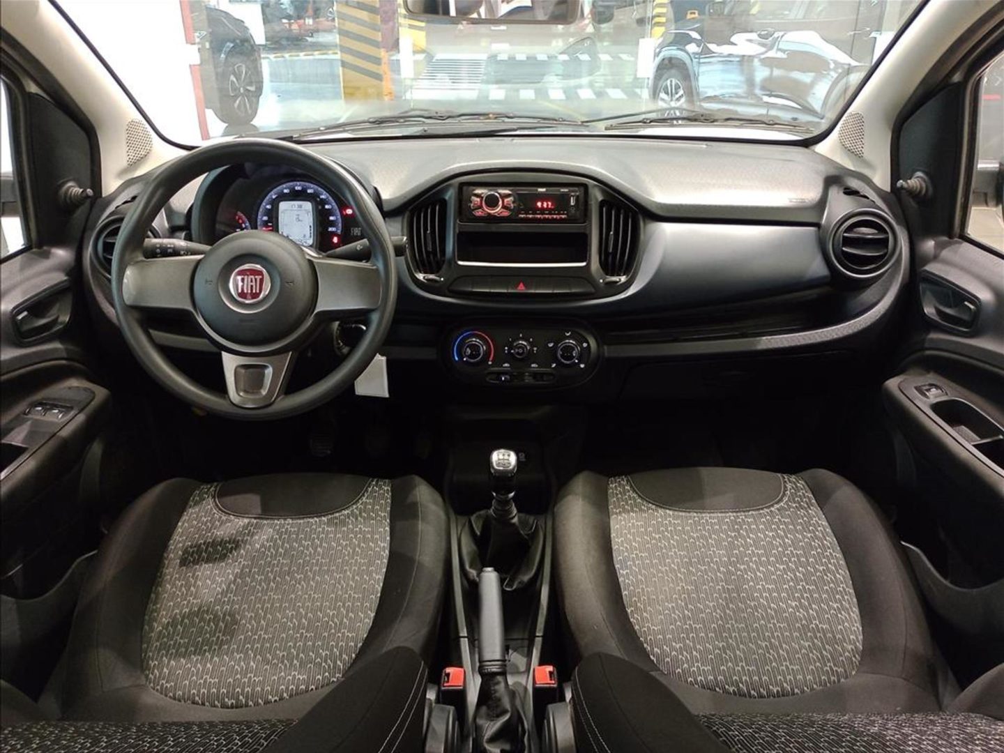 Foto interna do Fiat Uno Attractive 2021, exibindo painel de instrumentos, volante, parte dos bancos e portas.