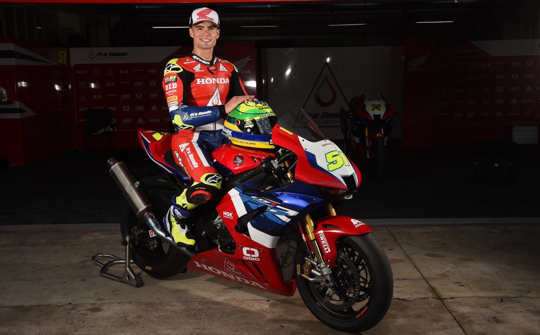 A imagem mostra o piloto de motociclismo Eric Granado em cima de sua moto de competição. Essa é uma foto posada. Ele está segurando o capacete nas cores verde, amarelo e azul, está vestindo um macacão de competição nas cores vermelha e azul, e sua moto está nas cores vermelha, azul e branca.