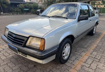 A imagem mostra um Chevrolet Monza na cor prata, estacionado na frente de um prédio.