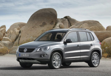 A imagem mostra um Volkswagen Tiguan G1, com pintura na cor prateada, parado na frente de rochas, em um ambiente com solo arenoso.