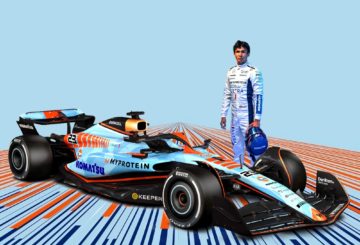 A imagem mostra o piloto Alexander Albon segurando um capacete e vestindo um mamacão da equipe Williams de F´pormula 1, ao lado de seu carro de corrida da categoria. O carro tem como cor predominante um azul claro e detalhes em laranja, que são as cores da empresa Gulf.