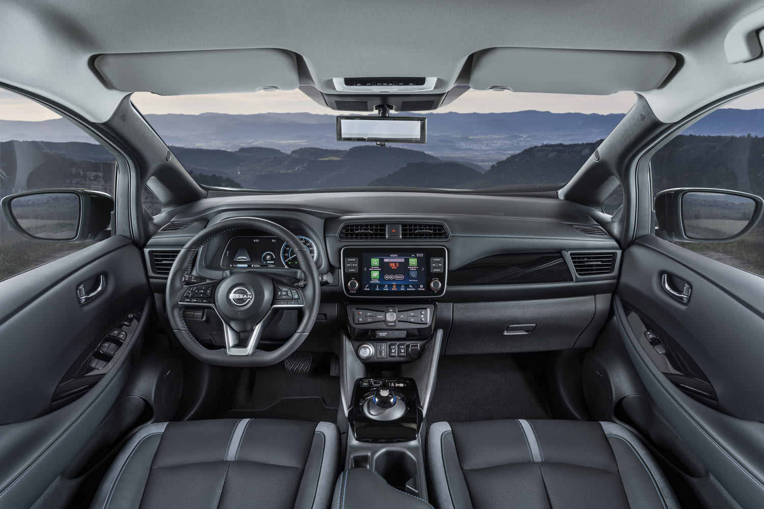Foto interna do Nissan Leaf 2024, exibindo o painel de instrumentos digital, volante multifuncional, central multimídia e parte dos bancos e portas do modelo.
