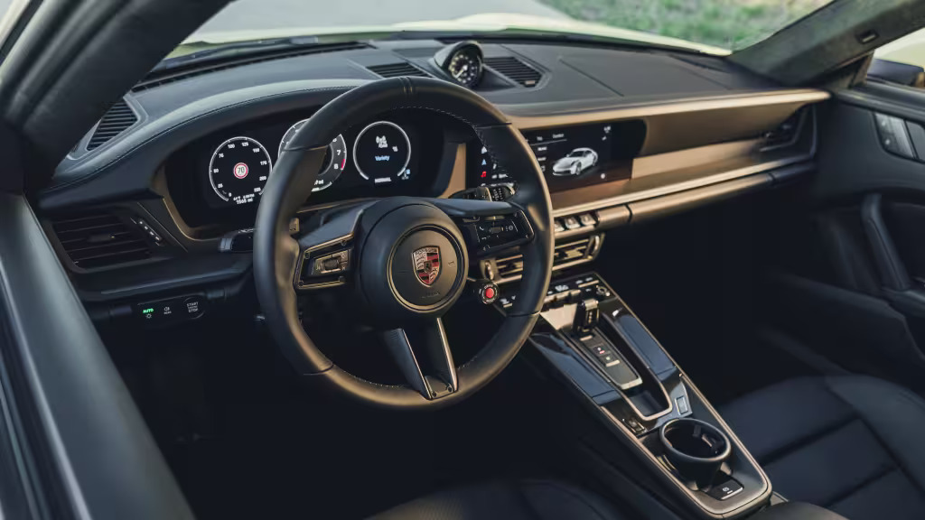 Foto interna do Porsche 911 Carrera S 2025, exibindo o painel de instrumentos, volante multifuncional, central multimídia e parte dos bancos.